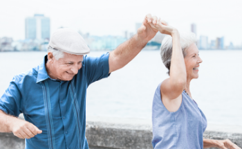 Older man and woman dancing and having fun outside near the sea with a city in the background.